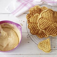 Runde Herzwaffeln aufeinander gestapelt auf einem Kuchengitter neben einer pinken Schüssel mit Waffelteig.