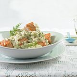 Schlotziges Risotto mit Lachs, Dill und Schnittlauch in einer weißen Schale auf einem Teller und weißer Tischdecke serviert.