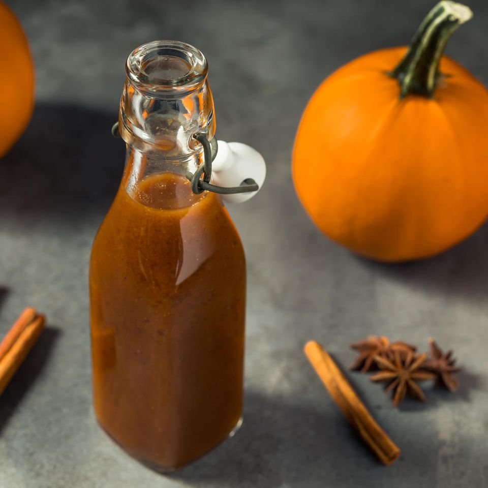 Selbst gemachter Pumpkin Spice Sirup in eine Flasche neben Zimtstangen, Anis, Nelken und Kürbissen auf grauem Untergrrund