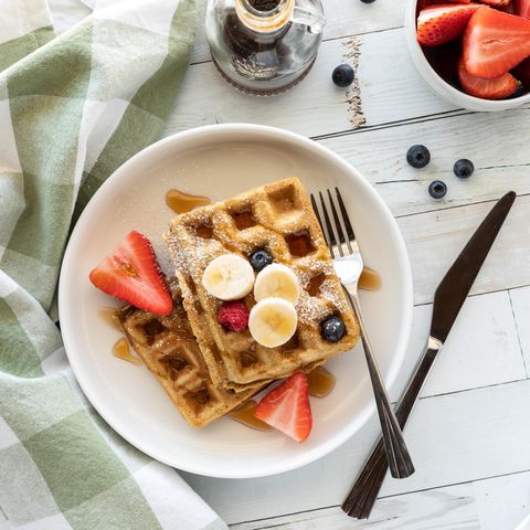 Zwei rechteckige Waffeln auf weoßem Teller mit Bananen, Erdbeeren, Heidelbeeren, Himbeere und Ahornsirup