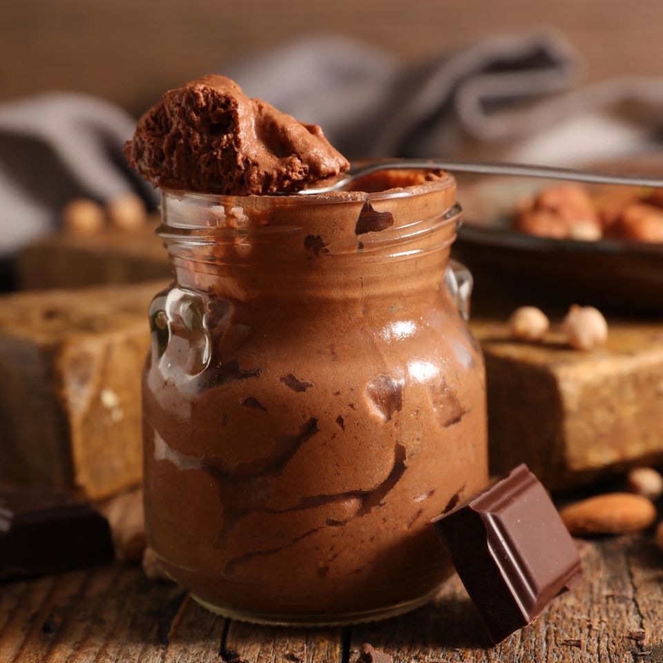 Vegane Mousse au Chocolat in einem Einmachglas. Ein Löffel mit Mousse liegt über der Glasöffnung, daneben Schokolade und Nüsse.