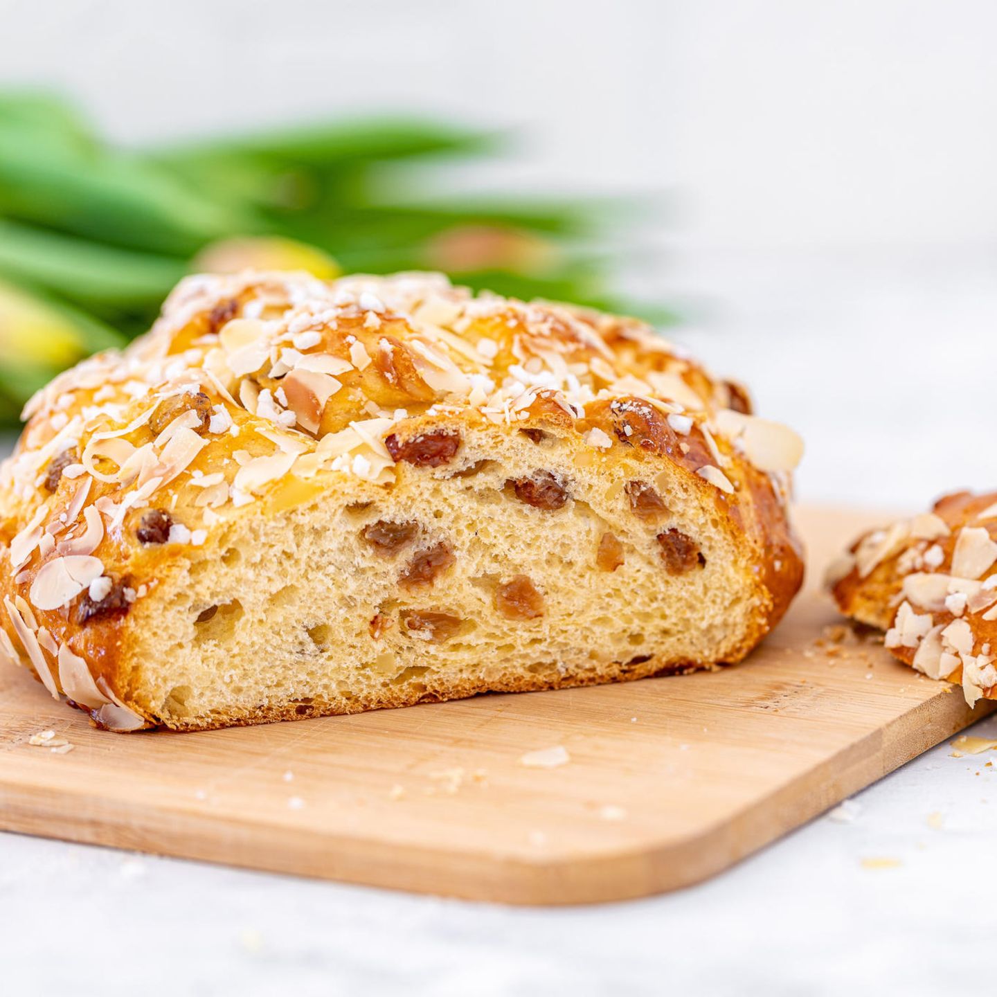 Angeschnittenes Osterbrot mit Rosinen, Hagelzucker und gehobelten Mandeln auf einem Holzbrett. Im Hintergrund Tulpen.