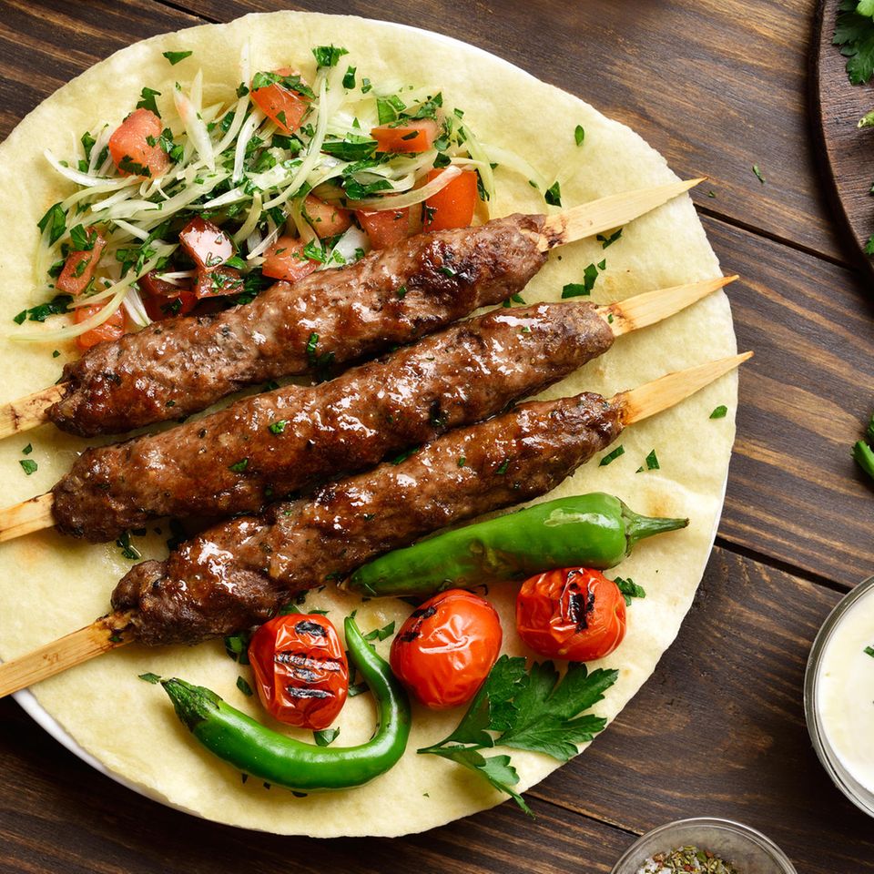 Türkische Hackfleischspieße auf einem Fladenbrot neben Zwiebelsalat mit Petersilie und Tomate und gegrilltem Gemüse.