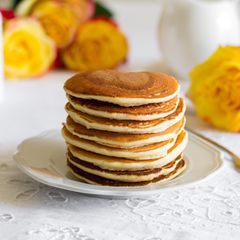 vegane Pancakes gestapelt auf weißem Teller und weißer Tischdecke mit Blumen im Hintergrund