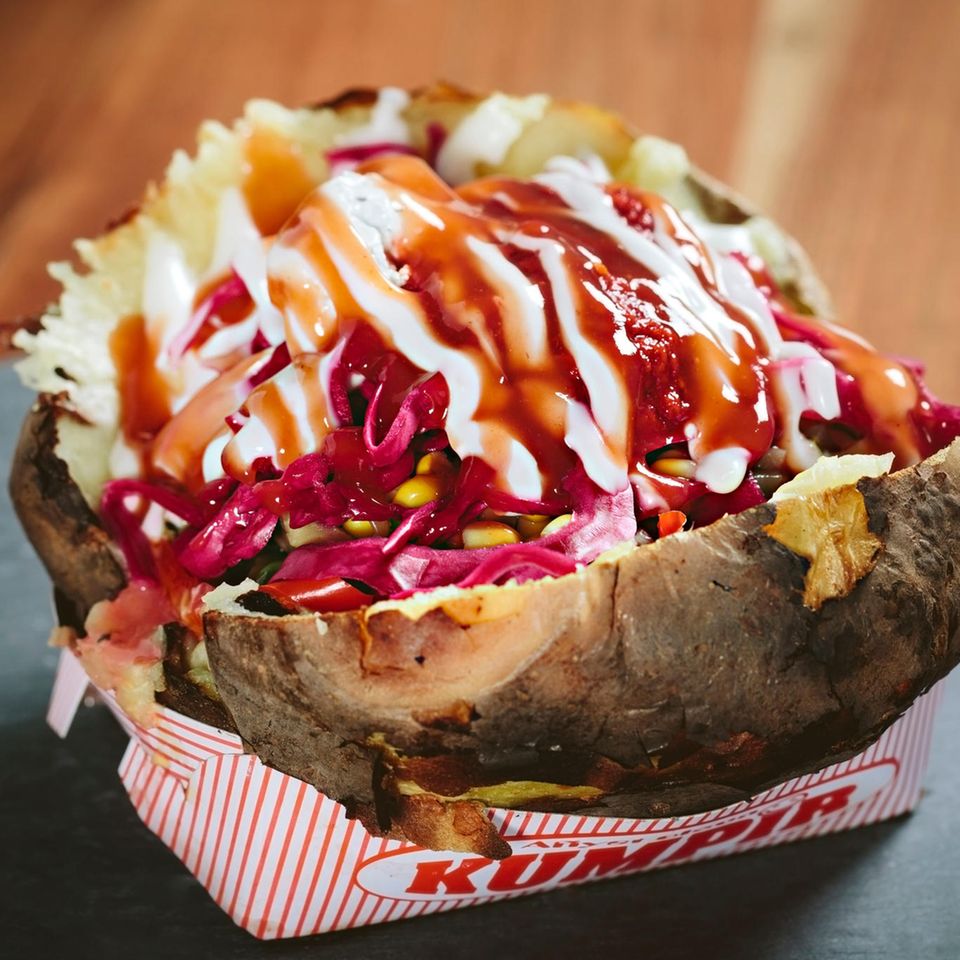 Gebackene Ofenkartoffel gefüllt mit Käse, Crème fraîche, Mais, Paprika, Frühlingszwiebel und Rotkraut in einem Papierschälchen.