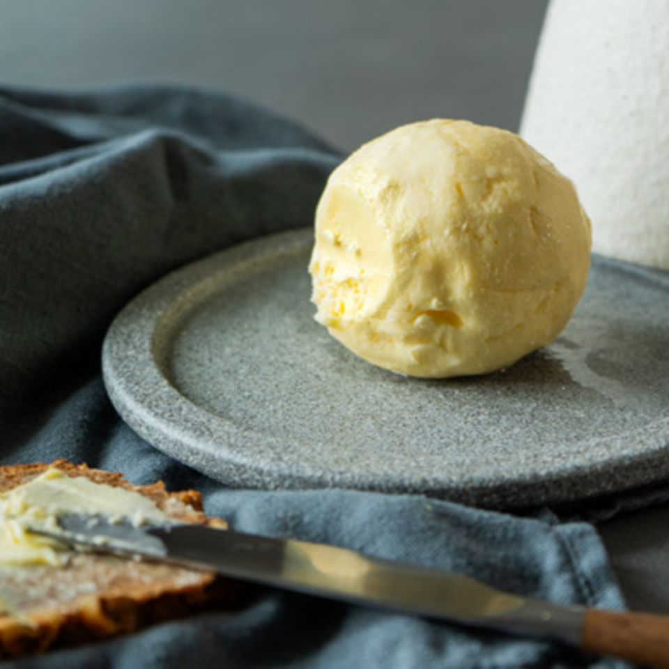 Selbst gemachte Butter zur Kugel geformt auf grauem Teller. Im Vordergrund eine Scheibe dunkles Brot mit Butter bestrichen.