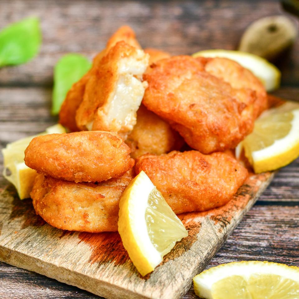 Knusprig frittierter Kibbeling auf einem Holzbrettchen serviert, mit Zitrone, Remoulade und Pommes im Hintergrund.