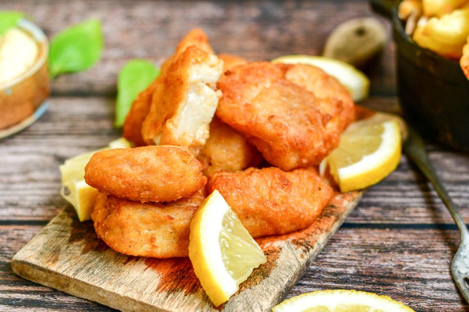 Knusprig frittierter Kibbeling auf einem Holzbrettchen serviert, mit Zitrone, Remoulade und Pommes im Hintergrund.