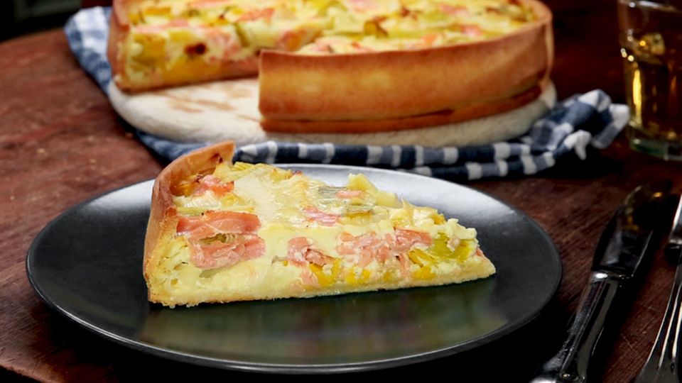 Eine Lachs-Quiche im Hintergrund, von der ein Stück herausgeschnitten wurde, welches vorne auf dem Teller liegt