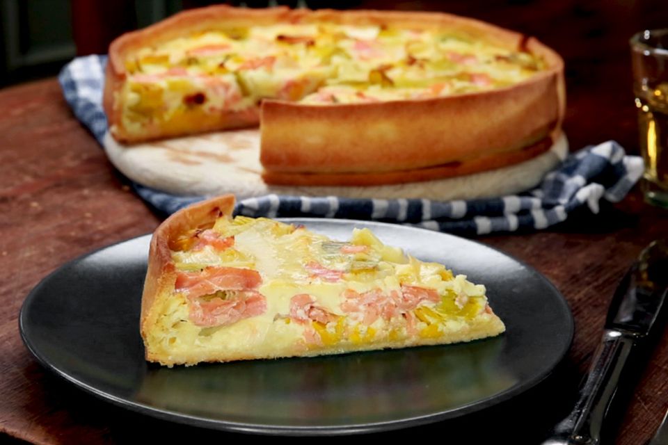 Eine Lachs-Quiche im Hintergrund, von der ein Stück herausgeschnitten wurde, welches vorne auf dem Teller liegt